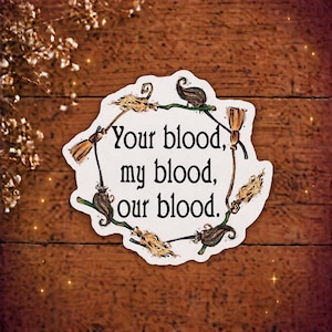 May include: A white die-cut sticker featuring a circular arrangement of six brown and green broomsticks surrounding the black text: Your blood, my blood, our blood. The sticker has a white border and is presented on a dark wooden surface.