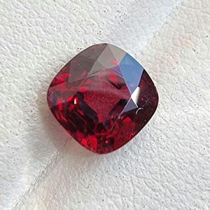 May include: A faceted, cushion-cut gemstone in a deep red colour. The gem's surface reflects light, showcasing its intricate internal structure. The stone is set against a white, textured background, highlighting its colour and clarity. This gem is suitable for jewellery.