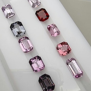 May include: Assortment of emerald-cut gemstones in various shades of pink, purple, and a deep red. The gems are arranged on a white surface, showcasing their facets and clarity. The collection includes light and dark hues.