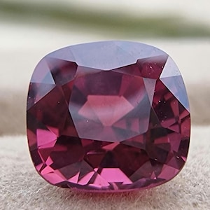 Puede incluir: Una piedra preciosa de color púrpura oscuro con un corte cojín. La piedra preciosa está facetada y tiene un alto pulido.