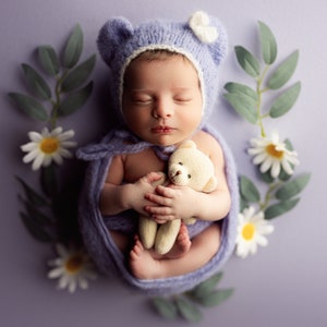 May include: A sleeping baby wearing a purple knitted hat with bear ears and holding a small teddy bear. The baby is surrounded by white daisies and green leaves on a purple background.