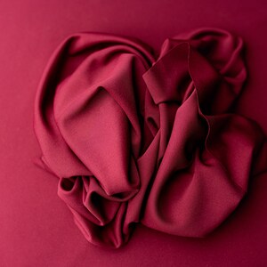 May include: A deep red fabric draped and folded in a heart shape. The fabric is smooth and silky.