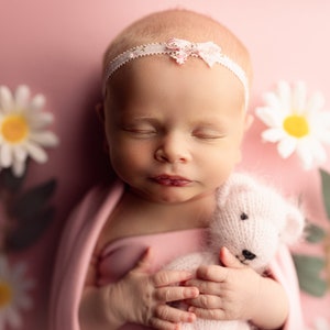 May include: A newborn baby lies on a pink surface, wrapped in a pink blanket, and holding a small, knitted teddy bear. The baby wears a white headband with a pink bow. White daisies and green leaves surround the baby.
