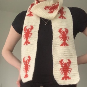May include: A white crocheted scarf with red lobster designs. The scarf is folded and worn around the neck of a person.