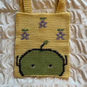 May include: Yellow crochet tote bag with a green alien face and purple fruit designs. The bag has two handles and is made of a soft, textured yarn. The alien face has black outlines and two small black eyes.