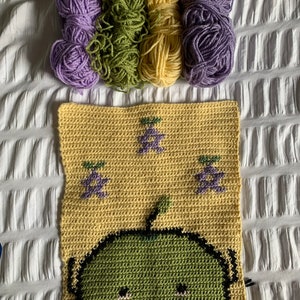 Puede incluir: Un proyecto de ganchillo con un cuadrado amarillo con un personaje de dibujos animados verde y dise&ntilde;os de uvas moradas. Varios ovillos de hilo en morado, verde y amarillo est&aacute;n dispuestos encima del cuadrado, sugiriendo un tema de manualidades.