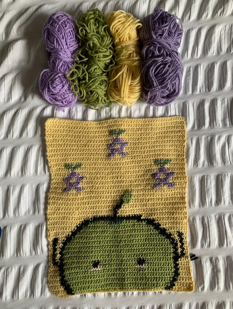 Puede incluir: Un proyecto de ganchillo con un cuadrado amarillo con un personaje de dibujos animados verde y dise&ntilde;os de uvas moradas. Varios ovillos de hilo en morado, verde y amarillo est&aacute;n dispuestos encima del cuadrado, sugiriendo un tema de manualidades.