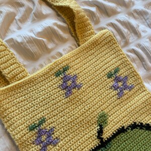 Puede incluir: Bolso tote de crochet amarillo hecho a mano con dos correas. El bolso presenta dise&ntilde;os florales morados y verdes bordados. El bolso est&aacute; hecho de un hilo suave y texturizado, adecuado para llevar varios art&iacute;culos.