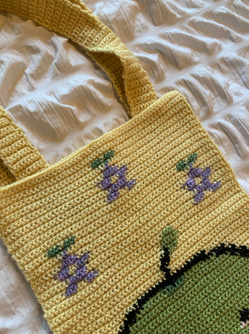 Puede incluir: Bolso tote de crochet amarillo hecho a mano con dos correas. El bolso presenta dise&ntilde;os florales morados y verdes bordados. El bolso est&aacute; hecho de un hilo suave y texturizado, adecuado para llevar varios art&iacute;culos.