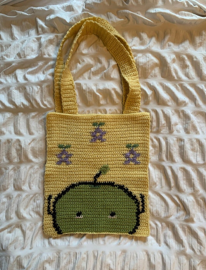 Puede incluir: Bolso tote de crochet amarillo con un dise&ntilde;o de cara de alien&iacute;gena verde y motivos de fruta morada. El bolso tiene dos asas y est&aacute; hecho de un hilo suave y texturizado. La cara del alien&iacute;gena tiene contornos negros y dos peque&ntilde;os ojos negros.