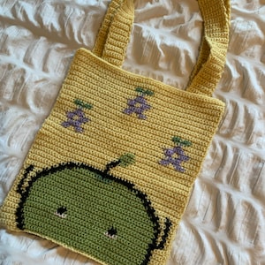 Puede incluir: Bolso tote de crochet hecho a mano en color amarillo claro. El bolso presenta un dise&ntilde;o de manzana verde en la parte delantera y racimos de uvas moradas. El bolso tiene dos asas amarillas y est&aacute; hecho de hilo suave.