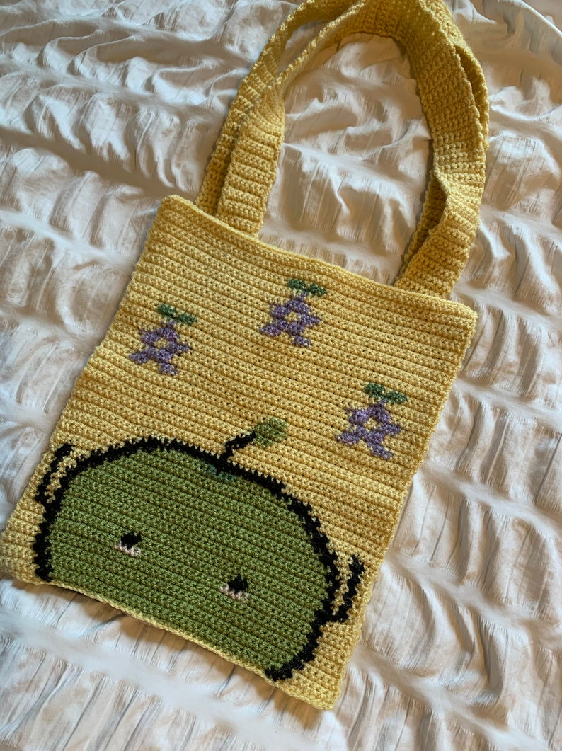 Puede incluir: Bolso tote de crochet hecho a mano en color amarillo claro. El bolso presenta un dise&ntilde;o de manzana verde en la parte delantera y racimos de uvas moradas. El bolso tiene dos asas amarillas y est&aacute; hecho de hilo suave.