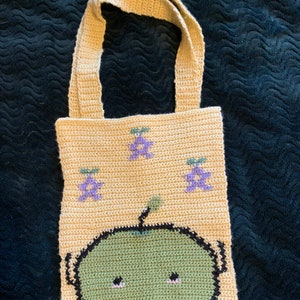 Puede incluir: Bolso tote de crochet beige con un dise&ntilde;o de cara de extraterrestre verde y detalles florales morados. El bolso tiene forma rectangular con dos asas beige. El extraterrestre tiene contornos negros, ojos negros peque&ntilde;os y un cuerpo verde.