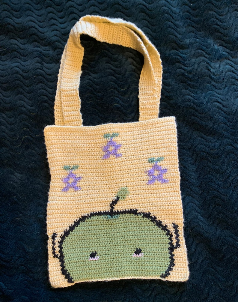 Puede incluir: Bolso tote de crochet beige con un dise&ntilde;o de cara de extraterrestre verde y detalles florales morados. El bolso tiene forma rectangular con dos asas beige. El extraterrestre tiene contornos negros, ojos negros peque&ntilde;os y un cuerpo verde.