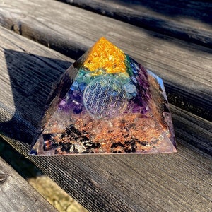 Op de afbeelding: Een zeven-chakra kristallen piramide met een levensbloem ontwerp in het midden. De piramide is gemaakt van helder hars en bevat verschillende kristallen, waaronder amethist, helder kwarts, citrien, groene aventurijn, lapis lazuli, tijgeroog en rode jaspis.