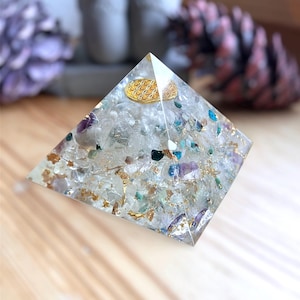 Può includere: Una piramide di cristallo trasparente con un simbolo dorato della fior di vita incastonato nella parte superiore. Il cristallo è pieno di piccoli pezzi di cristalli di vari colori, tra cui ametista, aventurina verde e quarzo chiaro.