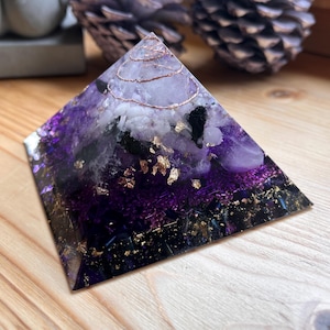 Könnte beinhalten: Eine handgefertigte Pyramiden-Skulptur mit einem geschichteten Design. Die Pyramide hat eine tiefviolette Basis, die in hellere Farbtöne mit Goldblattakzenten und einer Kupferdrahtspule an der Spitze übergeht. Ein einzigartiger Dekorationsartikel.