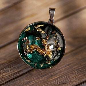 Op de afbeelding: Een ronde hangerketting met een groen en gouden harsontwerp. De hars heeft een draaiende structuur met gouden vlokken en een helder gedeelte in het midden.