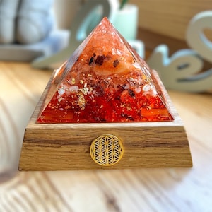 Puede incluir: Una pirámide de cristal roja con escamas doradas y otros cristales incrustados en su interior, sentada sobre una base de madera con un símbolo dorado de la flor de la vida.