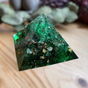 Puede incluir: Pirámide de resina verde con destellos dorados y cobrizos. La pirámide está hecha con resina transparente y tiene una variedad de destellos verdes, dorados y cobrizos.