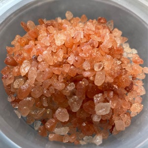 May include: A close-up view of a bowl filled with pink Himalayan salt crystals. The salt crystals are a mix of light and dark pink hues, with some white crystals visible.