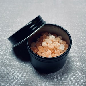 May include: A small black metal tin with a lid, filled with pink Himalayan salt crystals.