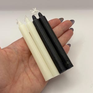 May include: Four taper candles, two ivory and two black, are held in a hand. The candles are slender and have a smooth finish. The wicks are visible at the top of each candle. The background is a plain white surface.