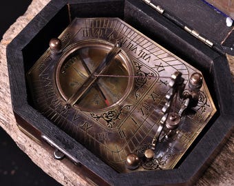 May include: An antique brass compass and sundial set in a dark wooden box. The compass features Roman numerals and intricate detailing. The box is open, revealing the compass and sundial, with a small latch. The compass is set on a wooden surface.