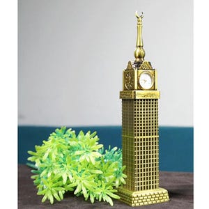 May include: A gold-colored metal model of the Abraj Al-Bait Clock Tower in Mecca, Saudi Arabia. The model features a working clock face and intricate details.