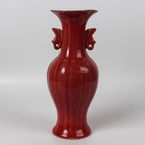 May include: A tall, red ceramic vase with a fluted body and two ornate handles. The vase has a flared rim and a glossy finish. The overall design is elegant and traditional, suitable for home decor.