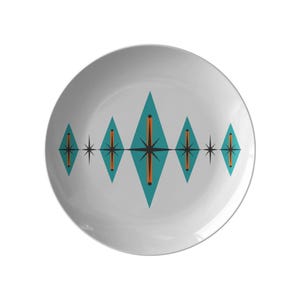 May include: A round white ceramic plate featuring a horizontal pattern of five teal diamond shapes. Each diamond contains a central black starburst design with a vertical orange line, interspersed with smaller black starburst accents.