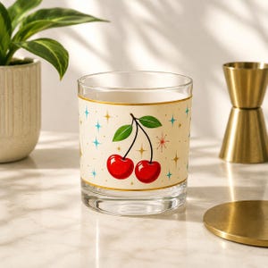 May include: A clear glass tumbler featuring a cream-colored band around the center. The band is decorated with two bright red cherries on green stems, surrounded by a pattern of small, multi-colored four-pointed stars and starburst shapes.