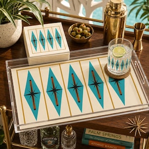 May include: A clear acrylic rectangular serving tray featuring a cream-colored insert with a repeating pattern of teal diamonds, gold vertical lines, and black starburst accents. A matching stack of paper napkins sits beside the tray.