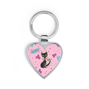 Könnte beinhalten: Herzförmiger Schlüsselanhänger mit silbernem Ring. Der rosa Hintergrund zeigt eine Illustration einer schwarzen Katze mit Weltraumthema. Die Katze hat Antennen und einen Kragen. Der Schlüsselanhänger hat den Text "Purr Baby".