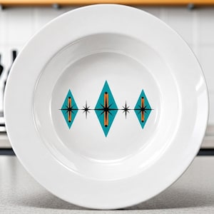 May include: A white ceramic bowl featuring a central graphic design of three teal diamond shapes. Each diamond contains a vertical orange line and a black starburst pattern. The design is symmetrical, with small black starbursts between the diamonds.