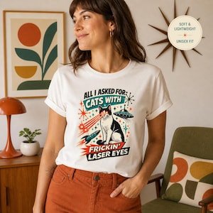 May include: White t-shirt with a graphic of a black and white cat with laser eyes, a rocket, and a UFO. Text reads "ALL I ASKED FOR: CATS WITH FRICKIN' LASER EYES." The shirt is labeled "SOFT & LIGHTWEIGHT" and "UNISEX FIT."