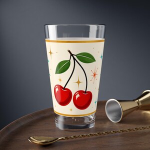 May include: A clear glass tumbler featuring a cream-colored band decorated with two bright red cherries on a single stem with green leaves, accented by small gold and teal starburst patterns against a plain background for a clean look.