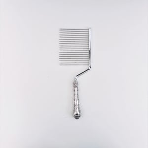 May include: A silver-toned cake slicer featuring a rectangular head with fifteen parallel thin metal wires attached to a decorative, ornate handle with embossed scrollwork details. The tool has a bent neck connecting the handle to the wire frame.