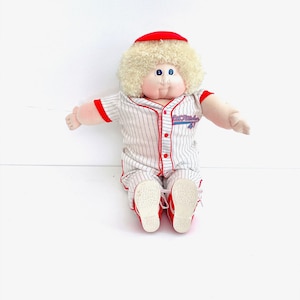 Könnte beinhalten: Eine Plüschpuppe mit lockigem blondem Haar und einer roten Mütze, bekleidet mit einer weißen Baseball-Uniform mit dünnen schwarzen Nadelstreifen, rotem Besatz und roten Knöpfen. Auf der Brust befindet sich der Schriftzug Cabbage Patch Kids.