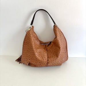 May include: A tan hobo bag featuring a textured ostrich-style leather finish with dark brown spots. The bag includes a single dark brown shoulder strap attached with silver-toned metal hardware and a decorative tassel detail on the side.