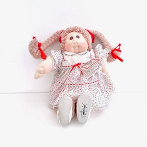 May include: A Cabbage Patch Kids doll with light brown yarn hair styled in two braids tied with red ribbons. The doll has a round face, a white dress with a small floral print, red trim, and lace edges. A tag is attached to the dress, and the doll wears white shoes with a signature on the sole.