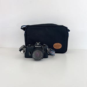 May include: A black Nikon film camera with a 50mm lens and a patterned camera strap, positioned in front of a black Vanguard camera bag. The camera features a silver shutter button, a lens cap, and metal strap lugs on both sides of the body.