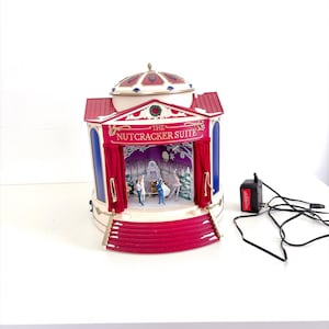 Puede incluir: Un expositor decorativo de teatro Nutcracker Suite con una estructura roja y blanca, techo abovedado, cortinas rojas y un escenario con figuras en miniatura. El letrero frontal dice THE NUTCRACKER SUITE. Incluye un adaptador de corriente negro.