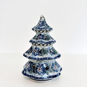 May include: A ceramic Christmas tree figurine featuring a hand-painted blue floral pattern on a white background. The tree has four tiers with cut-out star shapes and a decorative black lattice border detail along the edges of each branch layer.