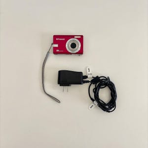 May include: A red Polaroid digital camera with a silver lens and a grey wrist strap, accompanied by a black power adapter with a coiled cord. The camera features the Polaroid logo and 7.0 megapixels text on the front face of the device.