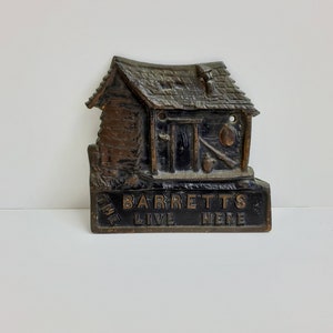 May include: A black metal sign with a raised image of a cabin and the text "The Barretts Live Here".