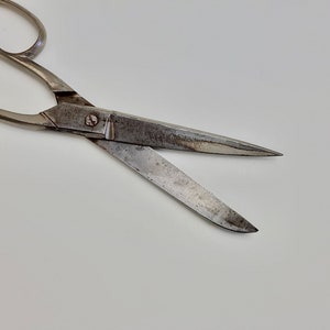 May include: Close-up of a pair of vintage scissors with silver handles and blades. The blades are open, revealing a worn, textured surface. The scissors are set against a plain white background.