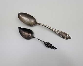 May include: Two antique silver spoons. One is a standard spoon with a floral design on the handle. The other is a small spoon with a leaf-shaped bowl and a floral design on the handle. Both spoons are on a white background.