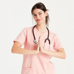 Könnte beinhalten: Ein rosa medizinischer Oberteil mit V-Ausschnitt und kurzen Ärmeln. Das Oberteil hat eine Tasche auf der linken Seite und ein Stethoskop um den Hals.
