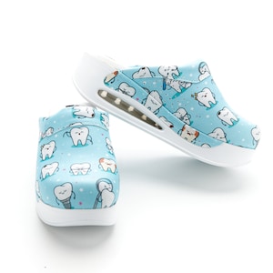 Puede incluir: Un par de zuecos de color azul claro y blanco con un patrón de dientes de dibujos animados. Los zuecos tienen una suela blanca y una parte superior azul claro con un ribete blanco. Los dientes de dibujos animados sonríen y tienen diferentes expresiones.